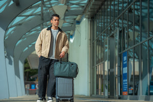 Traveler with INUK NAVI Black Forest Green backpack on a rolling suitcase, showing trolley sleeve use
